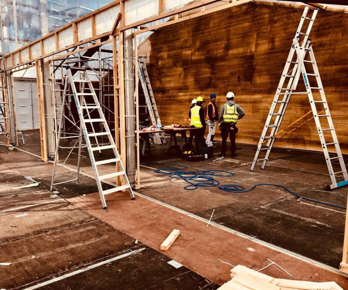 Construction scene with ladders, timber framework, and workers in safety gear assembling a large event structure with a wooden backdrop.