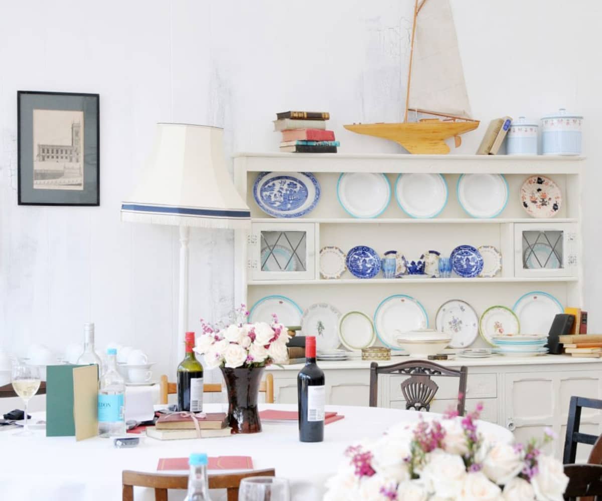  Elegant indoor dining setup at the Goodwood event, featuring a table with wine bottles, floral centrepieces, and a vintage dresser displaying decorative plates and nautical-themed ornaments.