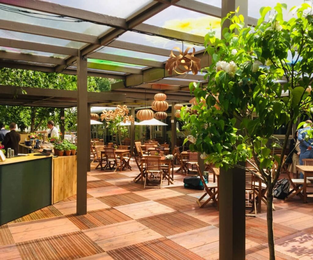 Open-air café space at the Royal Chelsea Flower Show with wooden decking, potted plants, hanging wooden light fixtures, and neatly arranged outdoor dining tables under a glass canopy.